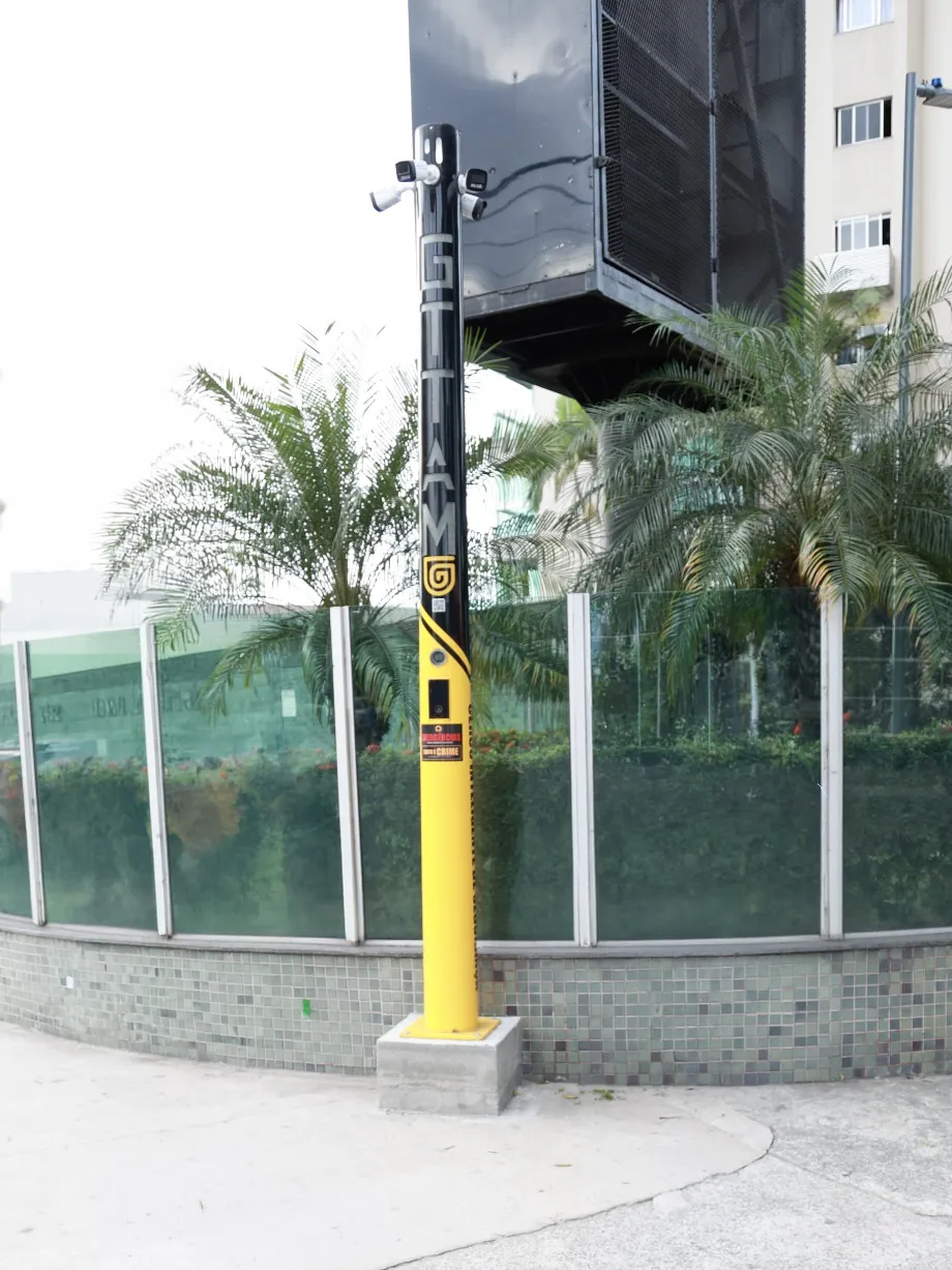 Torre de Monitoramento Inteligente da GTTAM instalada na calçada, com câmeras no topo, faixa preta com a marca e base amarela com videoporteiro